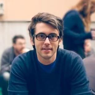 Sam Reinders, smiling slightly, wearing dark-rimmed glasses and a navy jumper, outdoors with people blurred in the background.